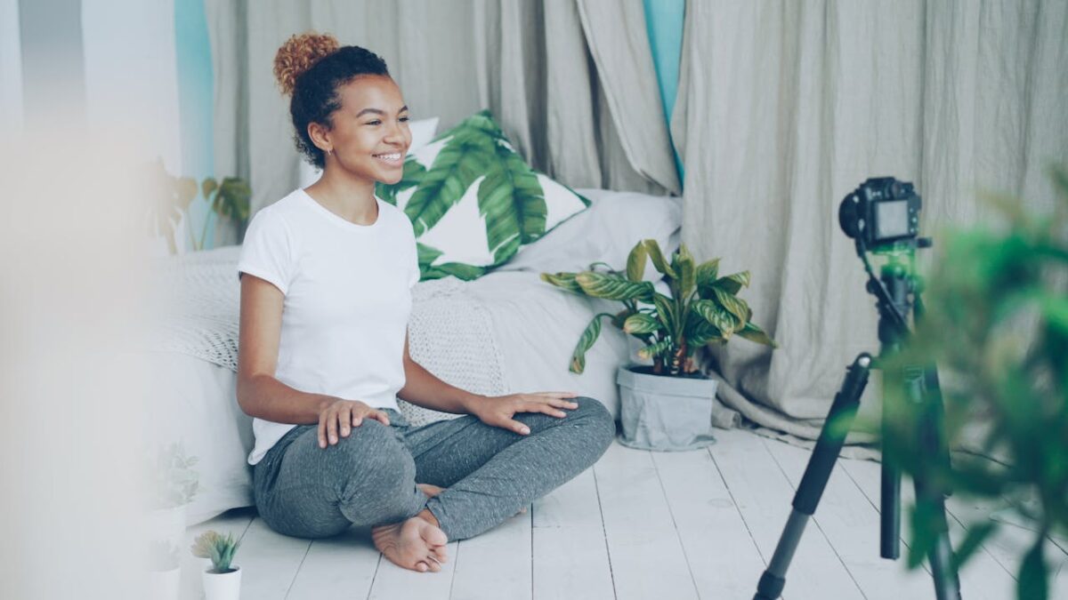 Smiling woman practicing yoga at home with camera setup, promoting wellness.