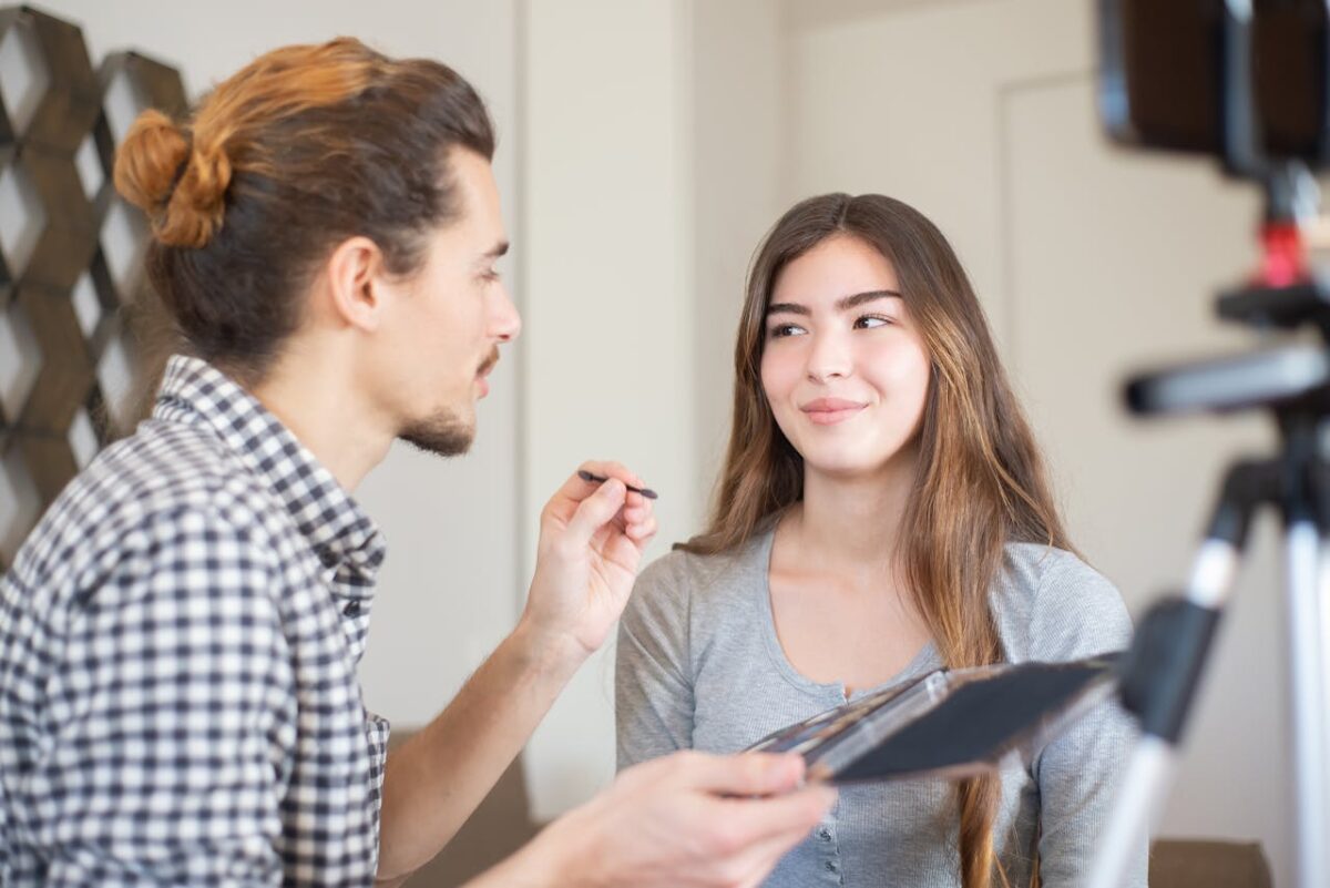 A makeup artist applies eyeshadow to a smiling woman during a vlog recording.